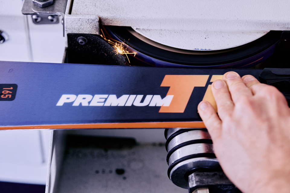 Eine Person schleift einen Ski mit der Aufschrift „PREMIUM T“ auf einer Schleifmaschine. In der Nähe der Schleifscheibe sind Funken sichtbar.