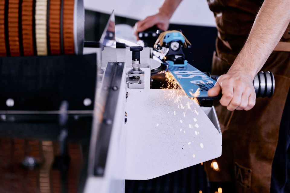 Eine Person schleift einen Ski auf einer Schleifmaschine. An der Kontaktstelle fliegen Funken. Die Person trägt eine Schürze und hält den Ski fest an der Maschine.