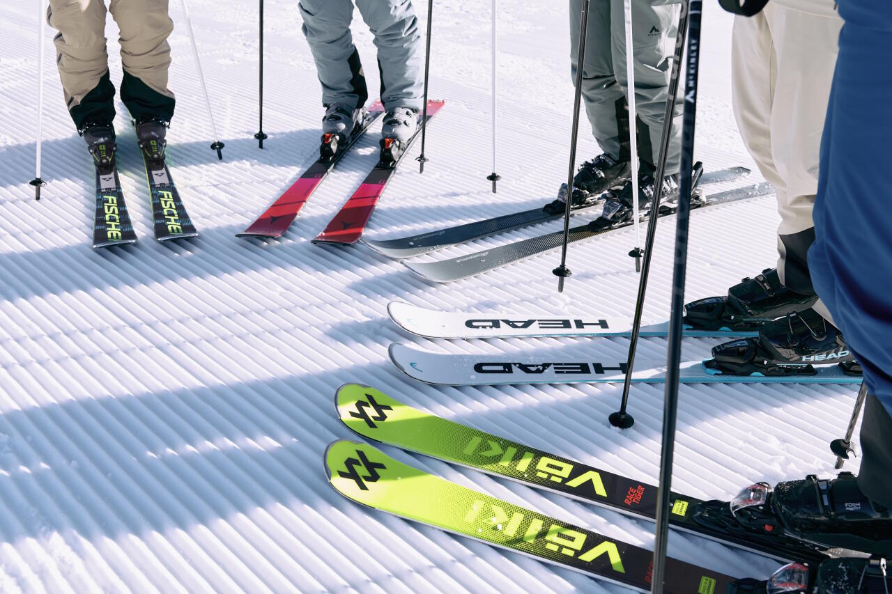 Mehrere Skifahrer stehen mit ihren bunten Skiern und Stöcken nebeneinander auf einer frisch präparierten Skipiste.