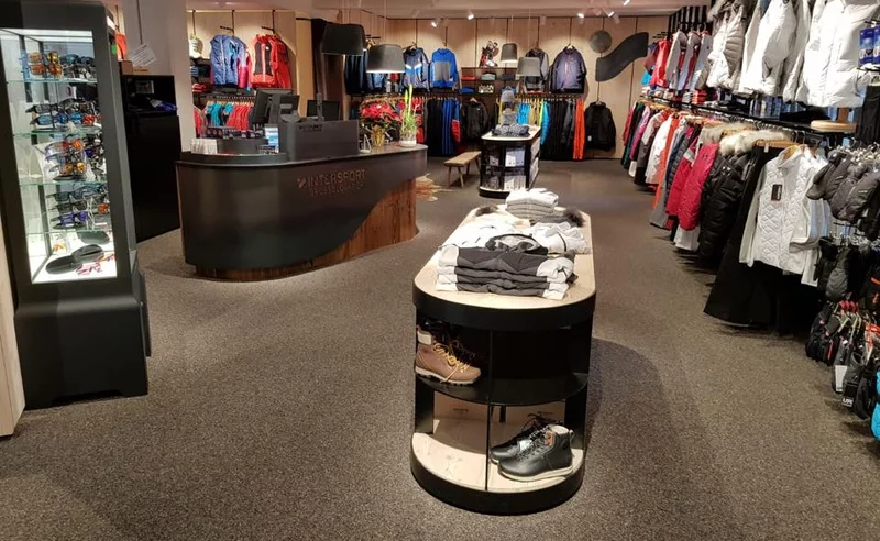 Ein Ladeninneres mit Regalen voller bunter Outdoor-Kleidung und Jacken auf beiden Seiten. In der Mitte befindet sich ein Ausstellungstisch mit gefalteter Kleidung und Schuhen. Das Geschäft ist modern gestaltet, mit Teppichboden und einer Theke im Hintergrund.