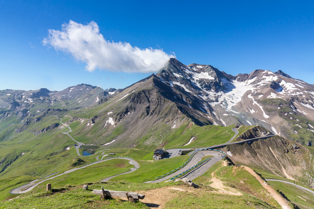 Ein malerischer Blick auf eine kurvenreiche Bergstraße, die durch üppig grüne Hügel zu einem schneebedeckten Gipfel unter einem klaren blauen Himmel führt, über dem eine einzelne Wolke schwebt.