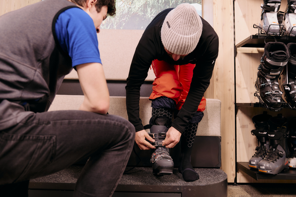 Eine Person mit grauer Mütze und roter Hose sitzt auf einer Bank und passt sich einen Skischuh an. Eine weitere Person in blauem Hemd und grauer Weste kniet davor und hilft beim Anpassen. Im Hintergrund sind Skischuhe in Regalen ausgestellt.