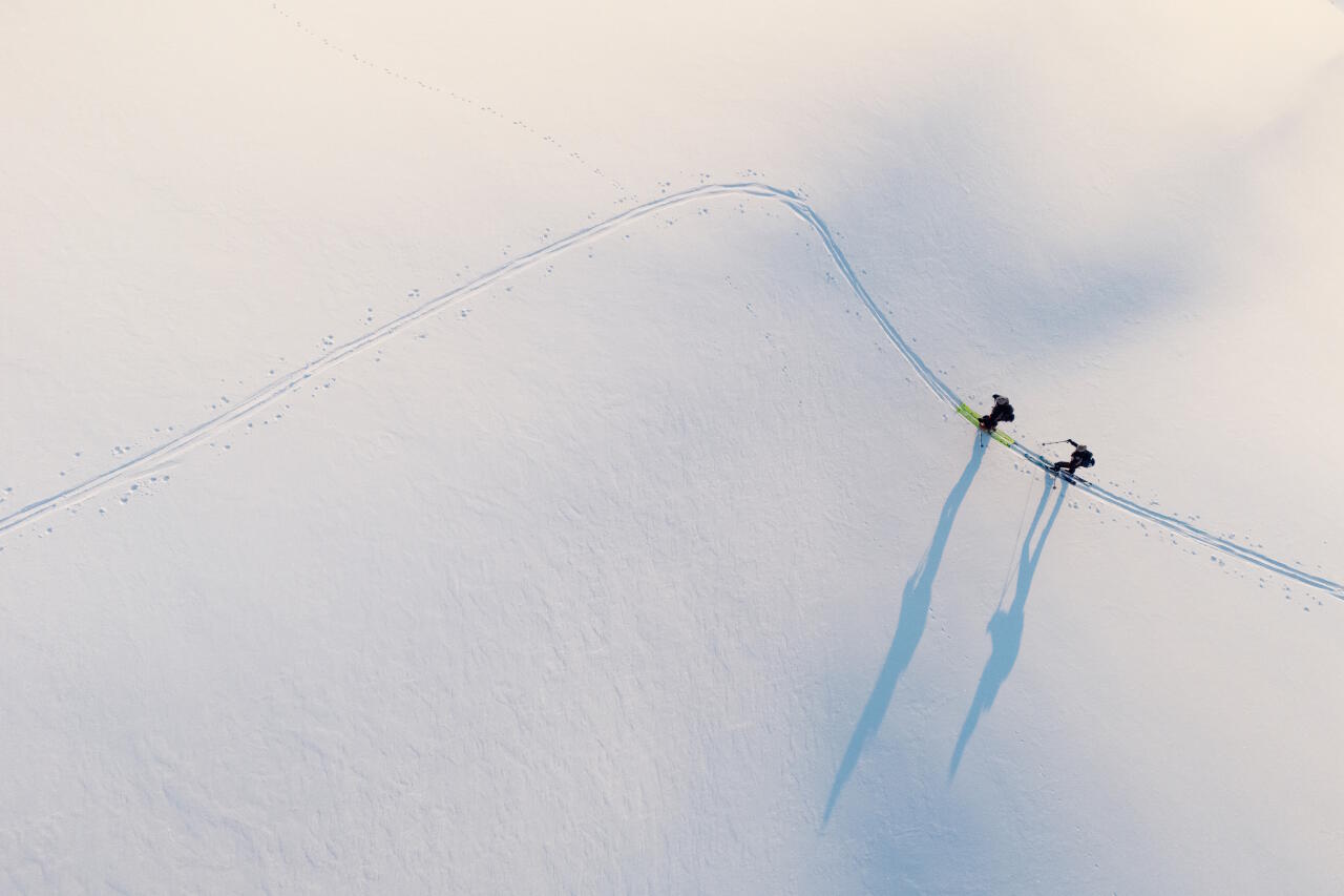 Zwei Skitourengeher steigen bei Sonnenlicht eine unberührte Schneefläche hinauf und werfen lange Schatten.