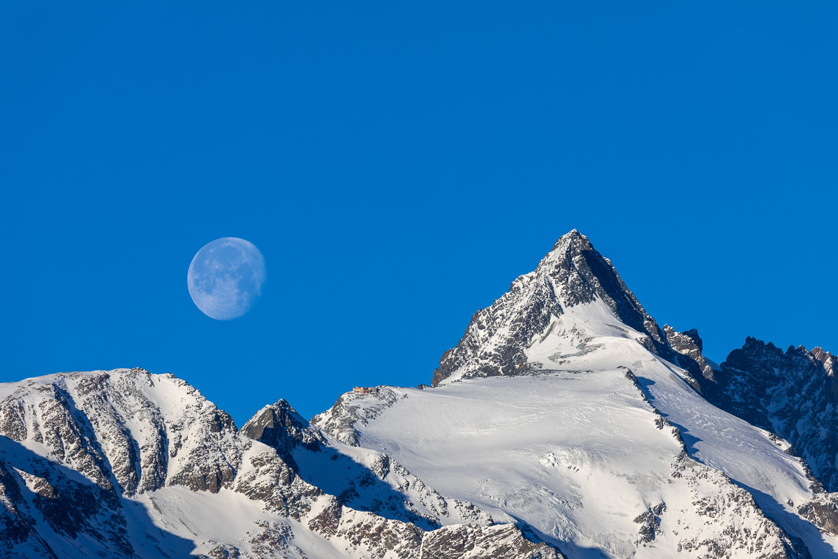 Ein schneebedeckter Berggipfel unter einem klaren blauen Himmel, links ist der Mond sichtbar.