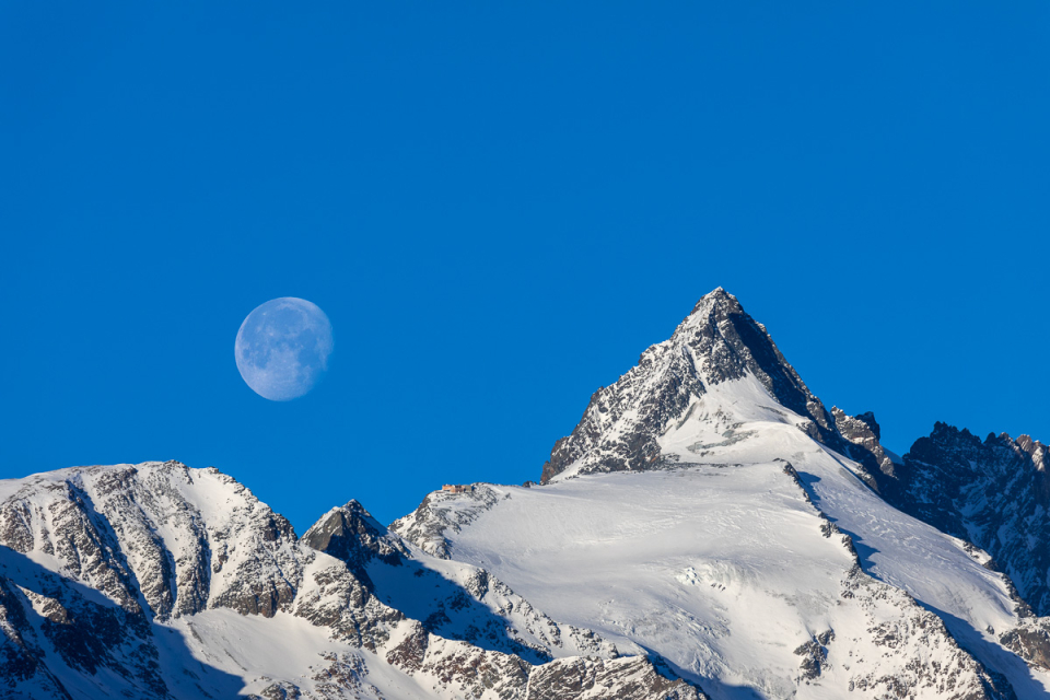 Ein schneebedeckter Berggipfel unter einem klaren blauen Himmel, links ist der Mond sichtbar.