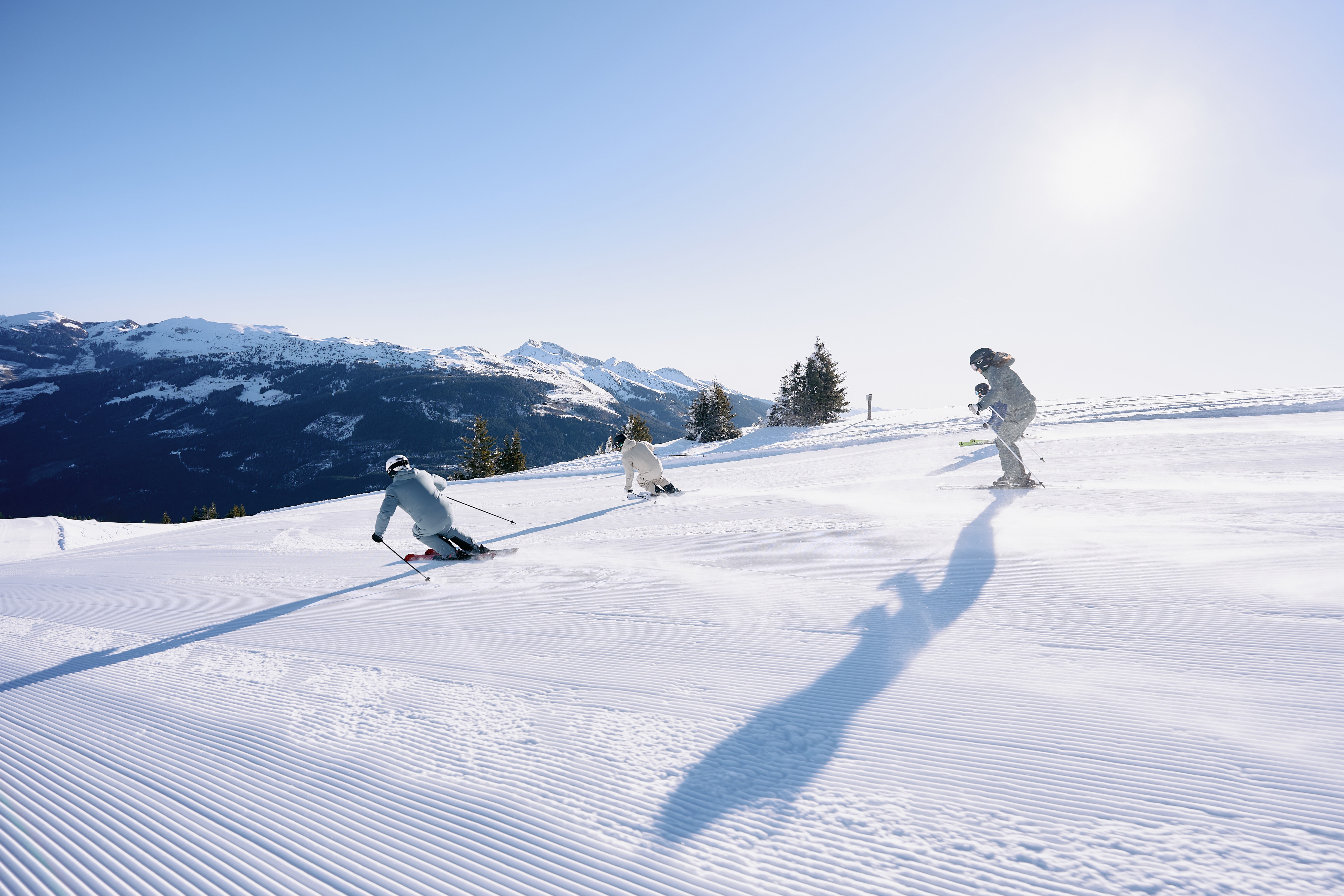 Drei Skifahrer fahren bei Sonnenschein eine frisch präparierte Skipiste mit Blick auf verschneite Berge hinab.