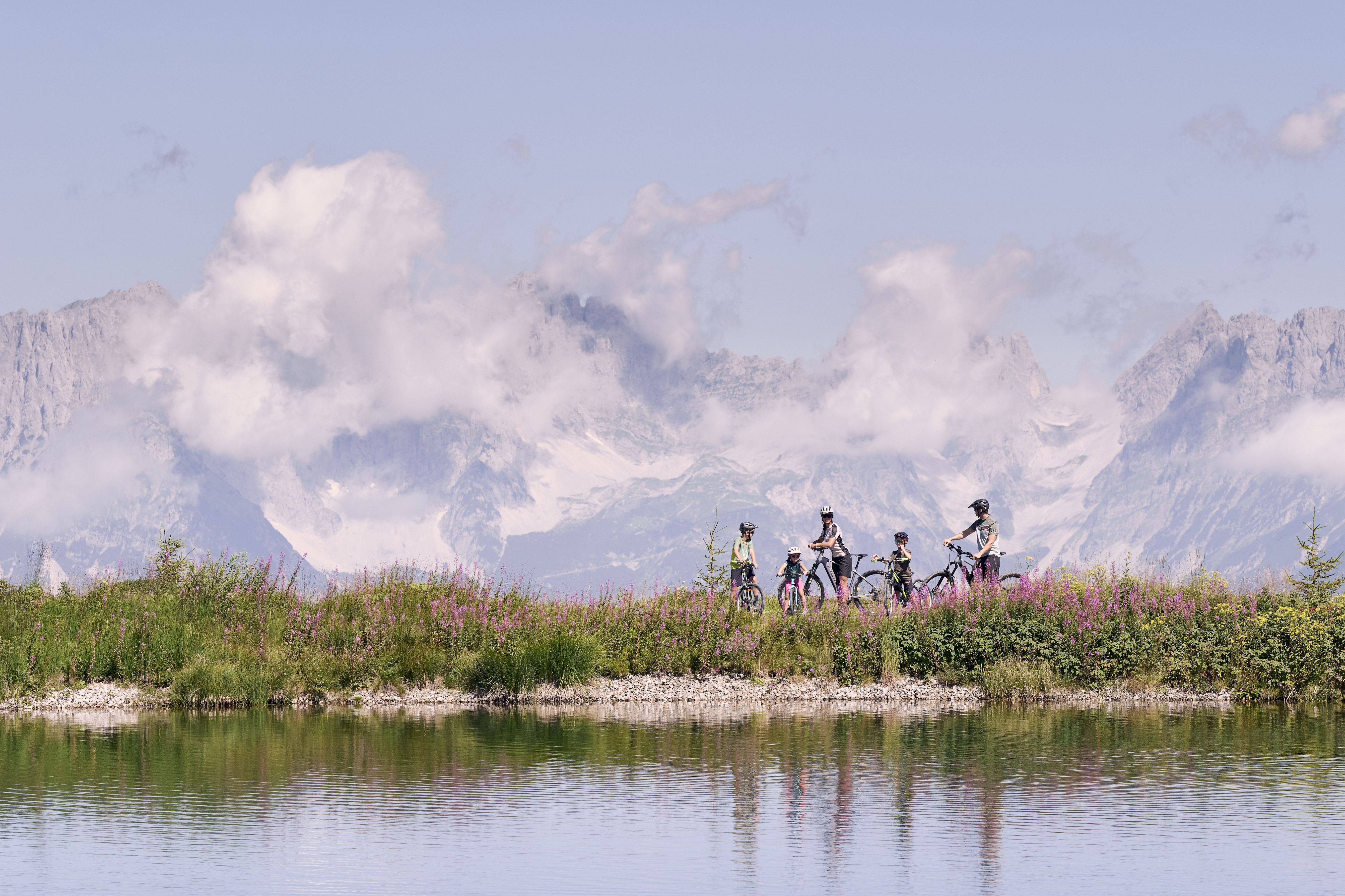 Eine Gruppe von Menschen fährt mit dem Fahrrad auf einem grasbewachsenen, von rosa Blumen gesäumten Weg, mit einem ruhigen See im Vordergrund und majestätischen, wolkenbedeckten Bergen im Hintergrund.