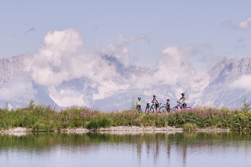A group of people riding bicycles on a grassy path lined with pink flowers, with a calm lake in the foreground and majestic, cloud-covered mountains in the background.