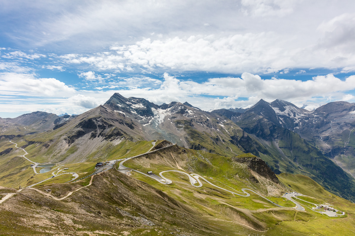 Eine malerische Aussicht auf eine Berglandschaft mit kurvenreichen Straßen, die sich durch grüne Hügel schlängeln. Im Hintergrund sind schneebedeckte Gipfel unter einem teilweise bewölkten Himmel zu sehen.