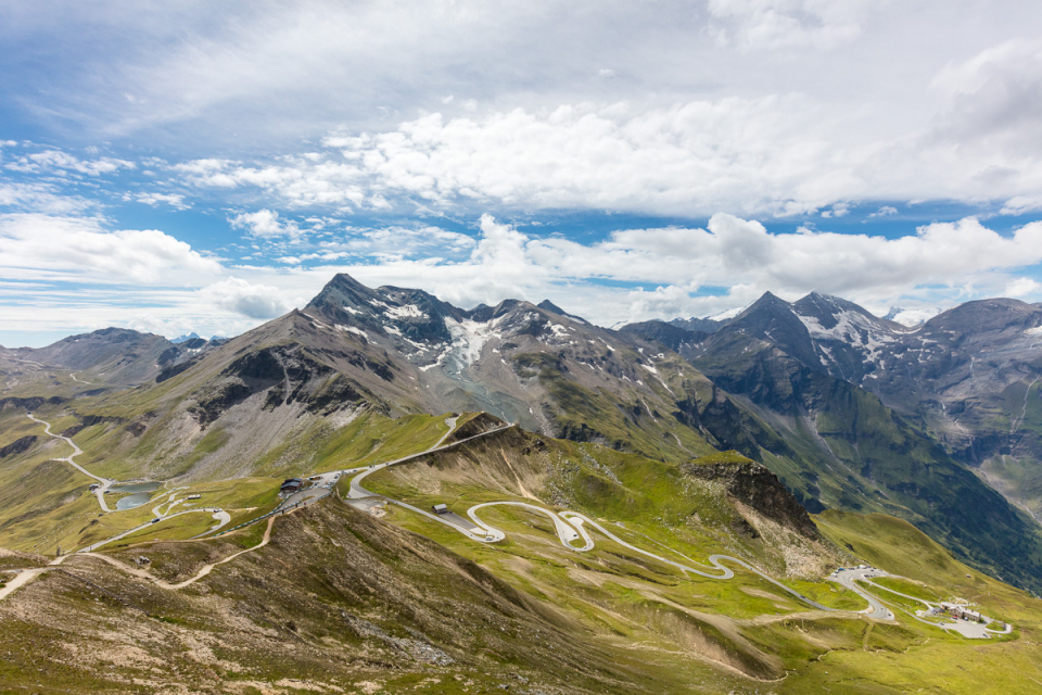 Eine malerische Aussicht auf eine Berglandschaft mit kurvenreichen Straßen, die sich durch grüne Hügel schlängeln. Im Hintergrund sind schneebedeckte Gipfel unter einem teilweise bewölkten Himmel zu sehen.