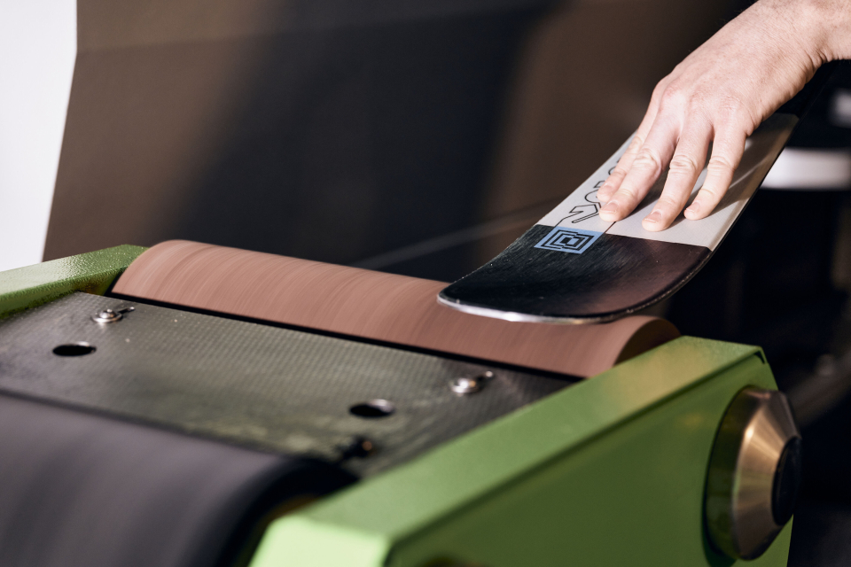 Eine Person schärft mit einer Maschine die Kante eines Skis. Der Ski liegt auf einem rotierenden Schleifband und wird von der Hand der Person geführt. Die Maschine ist grün und hat eine strukturierte Oberfläche.