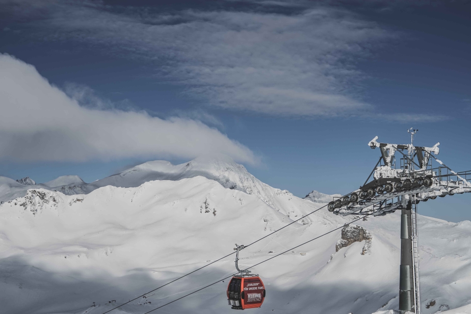 Vor der Kulisse schneebedeckter Berge und eines teilweise bewölkten Himmels fährt eine rote Seilbahn an einem Kabel entlang.