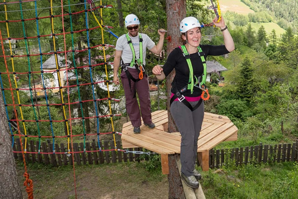 Two people wearing helmets and harnesses navigate a treetop adventure course. One person stands on a wooden platform attached to a tree, while the other is on a rope bridge. The background features trees and a view of a valley.