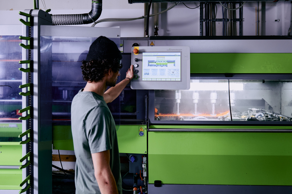 Eine Person mit Mütze und T-Shirt bedient eine große grüne Industriemaschine über einen Touchscreen. Die Maschine verfügt über einen transparenten Bereich, der die inneren Komponenten zeigt, und befindet sich in einer Werkstattumgebung.