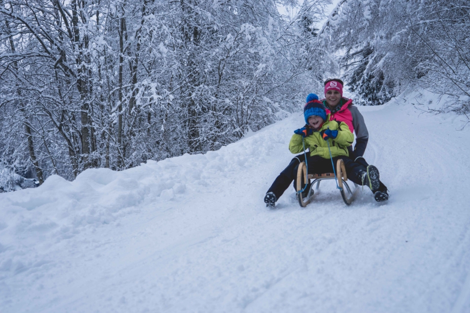 Ein lachendes Kind rodelt mit einem Erwachsenen auf einem Holzschlitten durch einen verschneiten Winterwald.