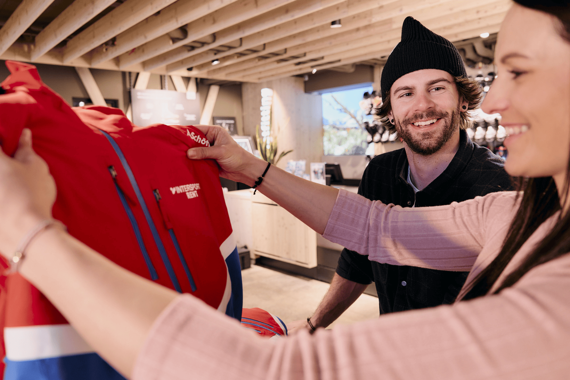 Eine Frau und ein Mann stehen in einem Geschäft und betrachten eine rote Jacke mit blauen und weißen Akzenten. Die Frau hält die Jacke in der Hand, während der Mann mit einer schwarzen Mütze sie anlächelt. Das Geschäft ist modern eingerichtet, mit Holzbalken und verschiedenen Produktpräsentationen im Hintergrund.