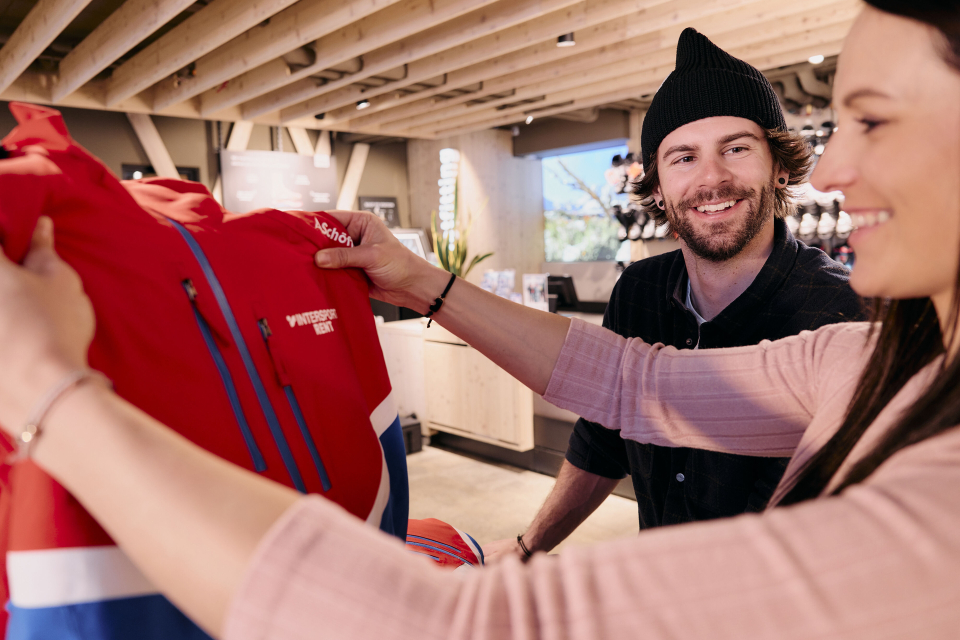 Eine Frau und ein Mann stehen in einem Geschäft und betrachten eine rote Jacke mit blauen und weißen Akzenten. Die Frau hält die Jacke in der Hand, während der Mann mit einer schwarzen Mütze sie anlächelt. Das Geschäft ist modern eingerichtet, mit Holzbalken und verschiedenen Produktpräsentationen im Hintergrund.