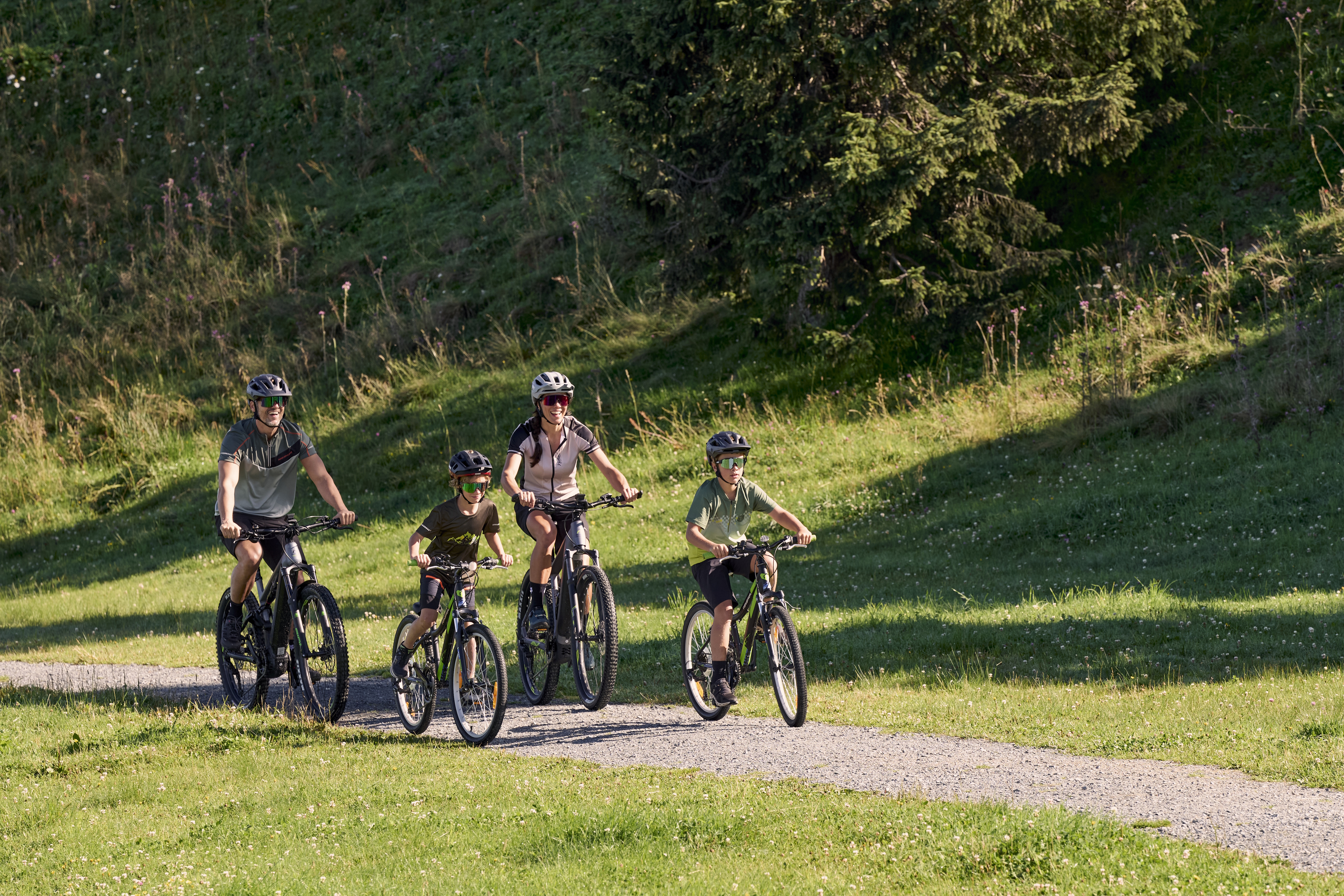 Eine vierköpfige Familie mit Helmen fährt gemeinsam mit dem Fahrrad auf einem asphaltierten Weg durch eine Rasenfläche mit Bäumen im Hintergrund. Die Sonne scheint und sorgt für eine helle und fröhliche Atmosphäre.