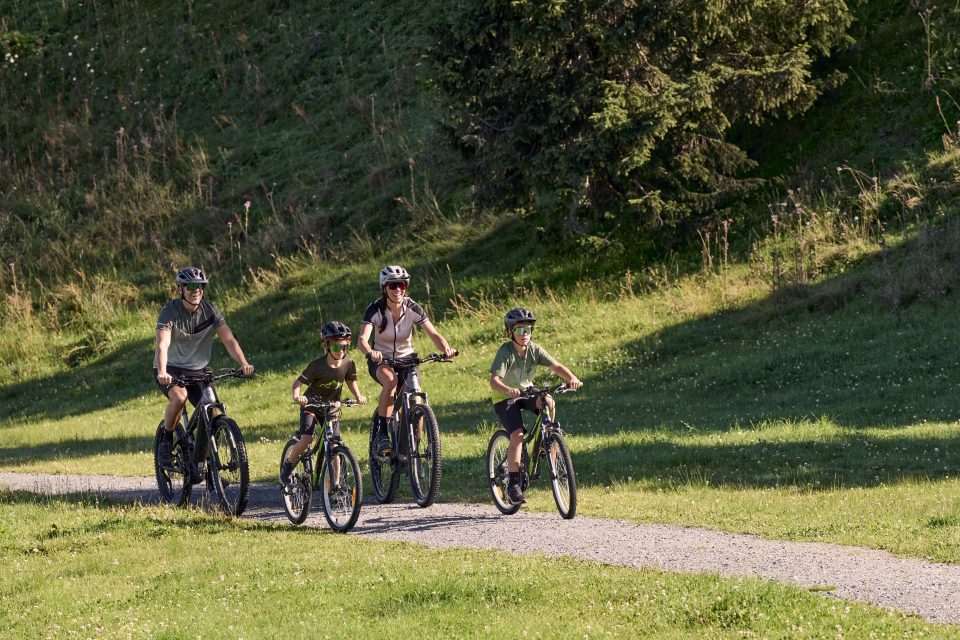 Eine vierköpfige Familie mit Helmen fährt gemeinsam mit dem Fahrrad auf einem asphaltierten Weg durch eine Rasenfläche mit Bäumen im Hintergrund. Die Sonne scheint und sorgt für eine helle und fröhliche Atmosphäre.