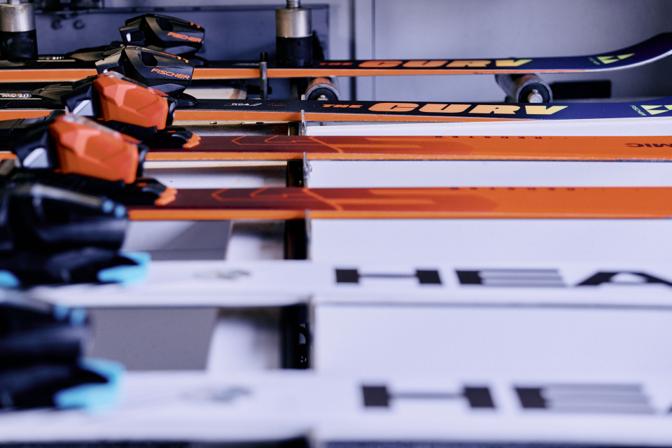 Eine Reihe horizontal aufgereihter Skier verschiedener Marken und Farben, darunter orange und weiße Skier mit sichtbaren Bindungen.