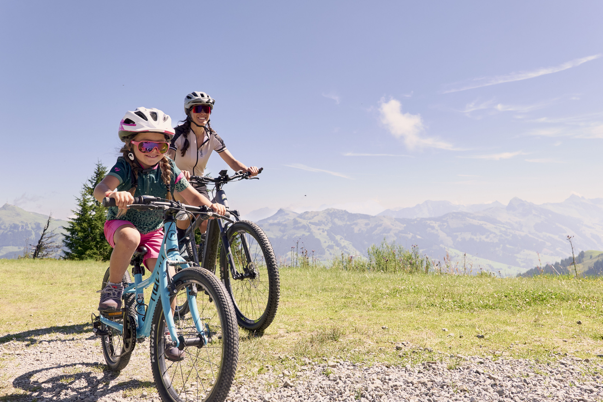 Ein Kind und ein Erwachsener fahren mit dem Fahrrad auf einem grasbewachsenen Weg mit einer malerischen Berglandschaft im Hintergrund. Beide tragen Helme und Sonnenbrillen und genießen einen sonnigen Tag im Freien.