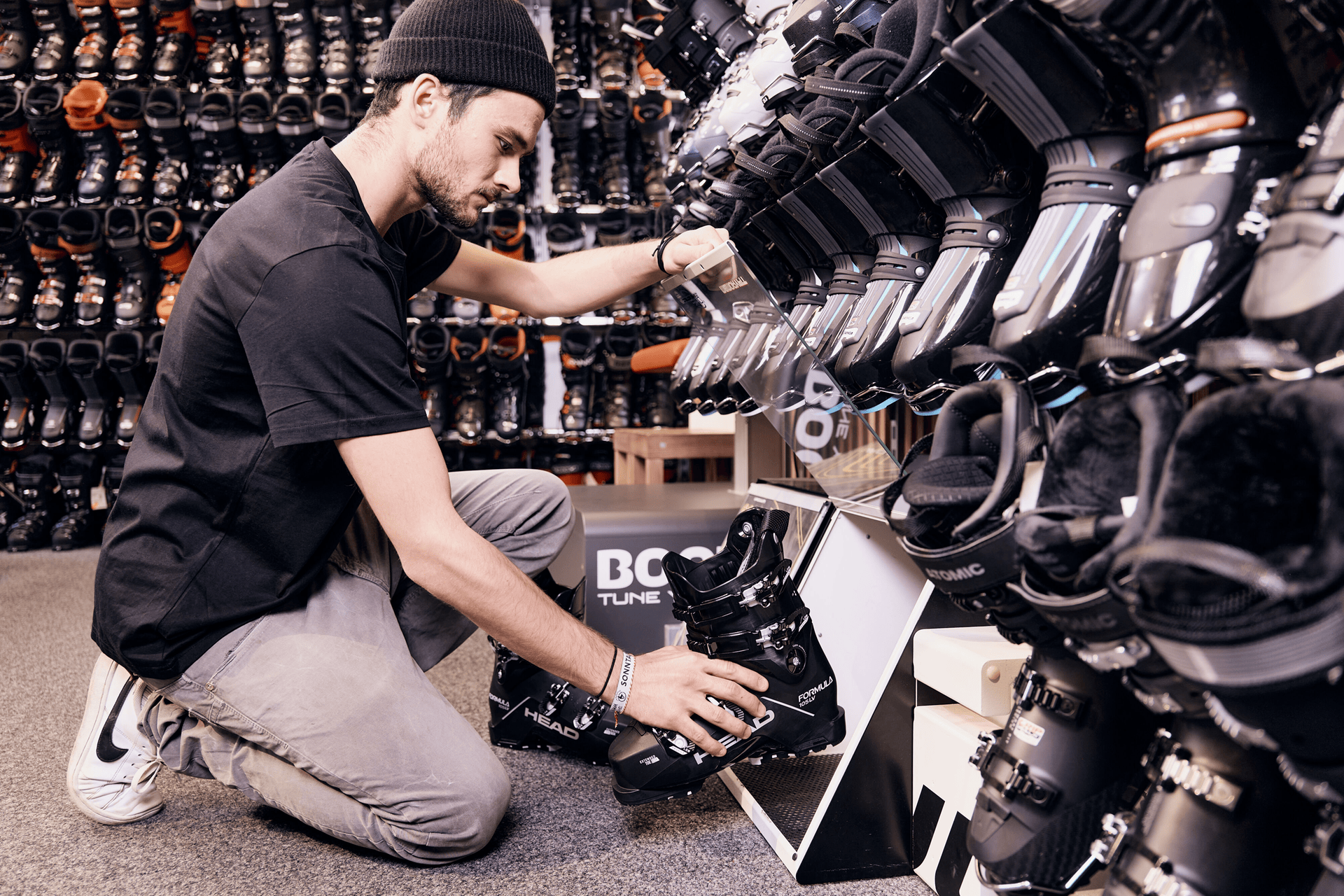 Eine Person mit schwarzer Mütze und schwarzem Hemd kniet in einem Geschäft auf dem Boden und betrachtet ein Paar schwarze Skischuhe in einem Ausstellungsregal. Im Hintergrund sind verschiedene Skischuhe in den Regalen angeordnet.