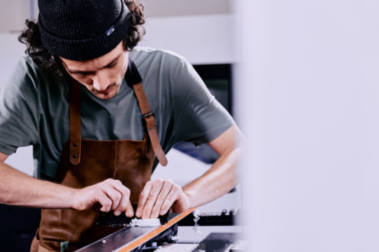 Eine Person mit Mütze und Schürze arbeitet konzentriert mit einem Werkzeug an einem Gerät, möglicherweise schärft oder justiert es. Die Szene scheint eine Werkstatt oder ein Studio zu sein.