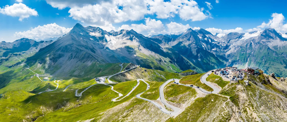 Ein Panoramablick auf eine Berglandschaft mit kurvenreichen Straßen, die sich durch grüne Hügel schlängeln. Im Hintergrund erheben sich schneebedeckte Gipfel unter einem teilweise bewölkten blauen Himmel. Auf einem der Hügel ist eine kleine Ansammlung von Gebäuden zu sehen.