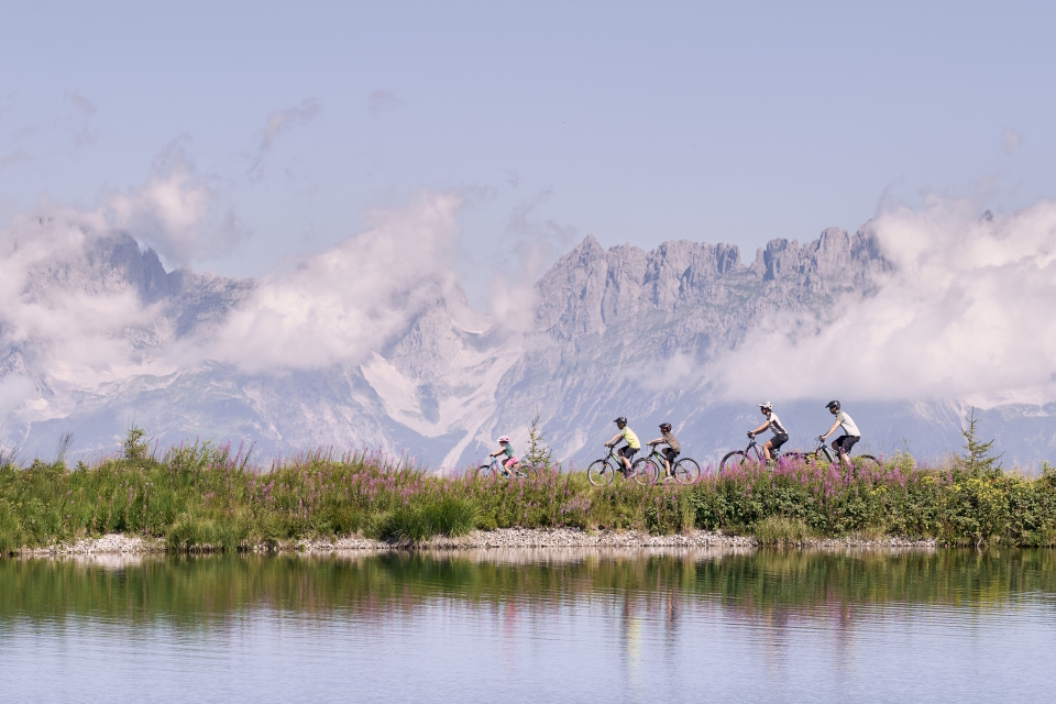 Eine Gruppe von fünf Personen radelt auf einem Weg neben einem ruhigen See entlang, im Hintergrund sind die Berge teilweise von Wolken bedeckt.