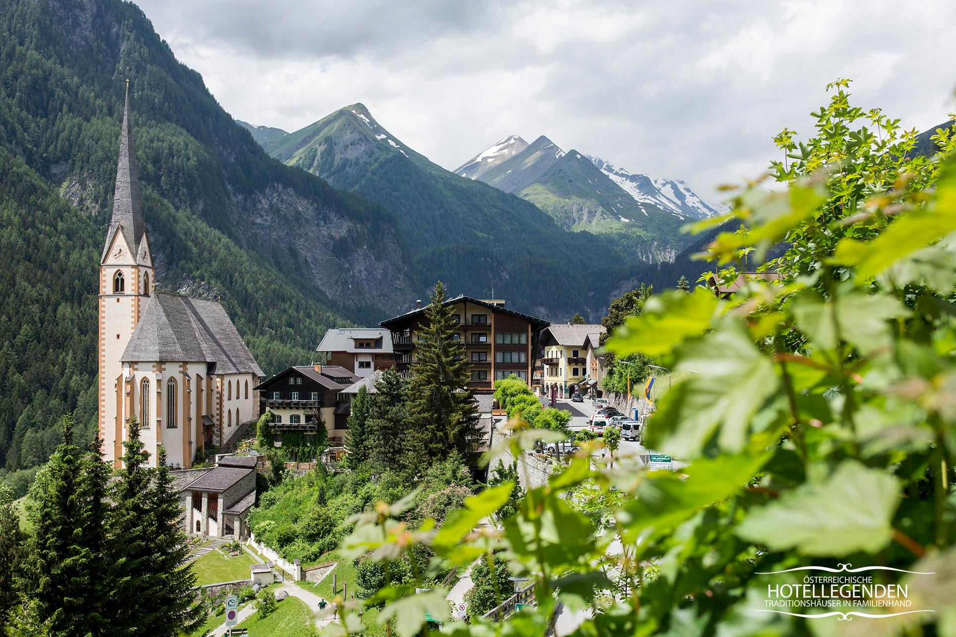 Ein malerisches Dorf inmitten einer Berglandschaft mit einer markanten Kirche mit hohem Turm. Rund um die Kirche befinden sich traditionelle alpine Gebäude, im Hintergrund üppiges Grün und hoch aufragende Berge.