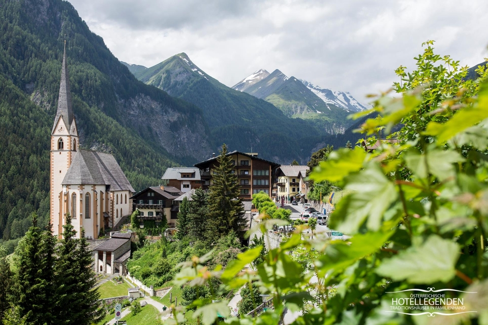 Ein malerisches Dorf inmitten einer Berglandschaft mit einer markanten Kirche mit hohem Turm. Rund um die Kirche befinden sich traditionelle alpine Gebäude, im Hintergrund üppiges Grün und hoch aufragende Berge.