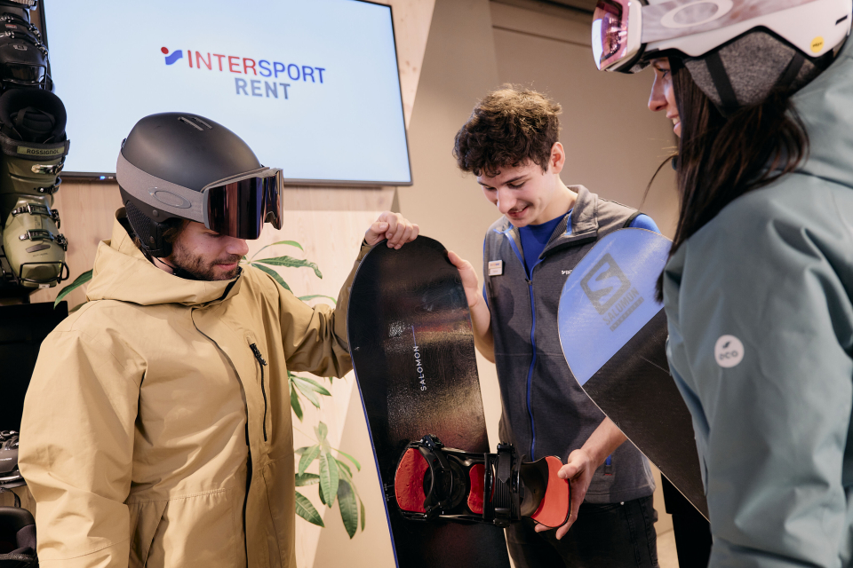 Drei Personen in einem Skiverleih begutachten Snowboards. Eine Person in beiger Jacke und Helm hält ein Snowboard, eine andere Person in blauer Jacke hilft. Eine dritte Person mit Helm hält ein weiteres Snowboard. Im Hintergrund ist ein Schild mit der Aufschrift „INTERSPORT RENT“ zu sehen.