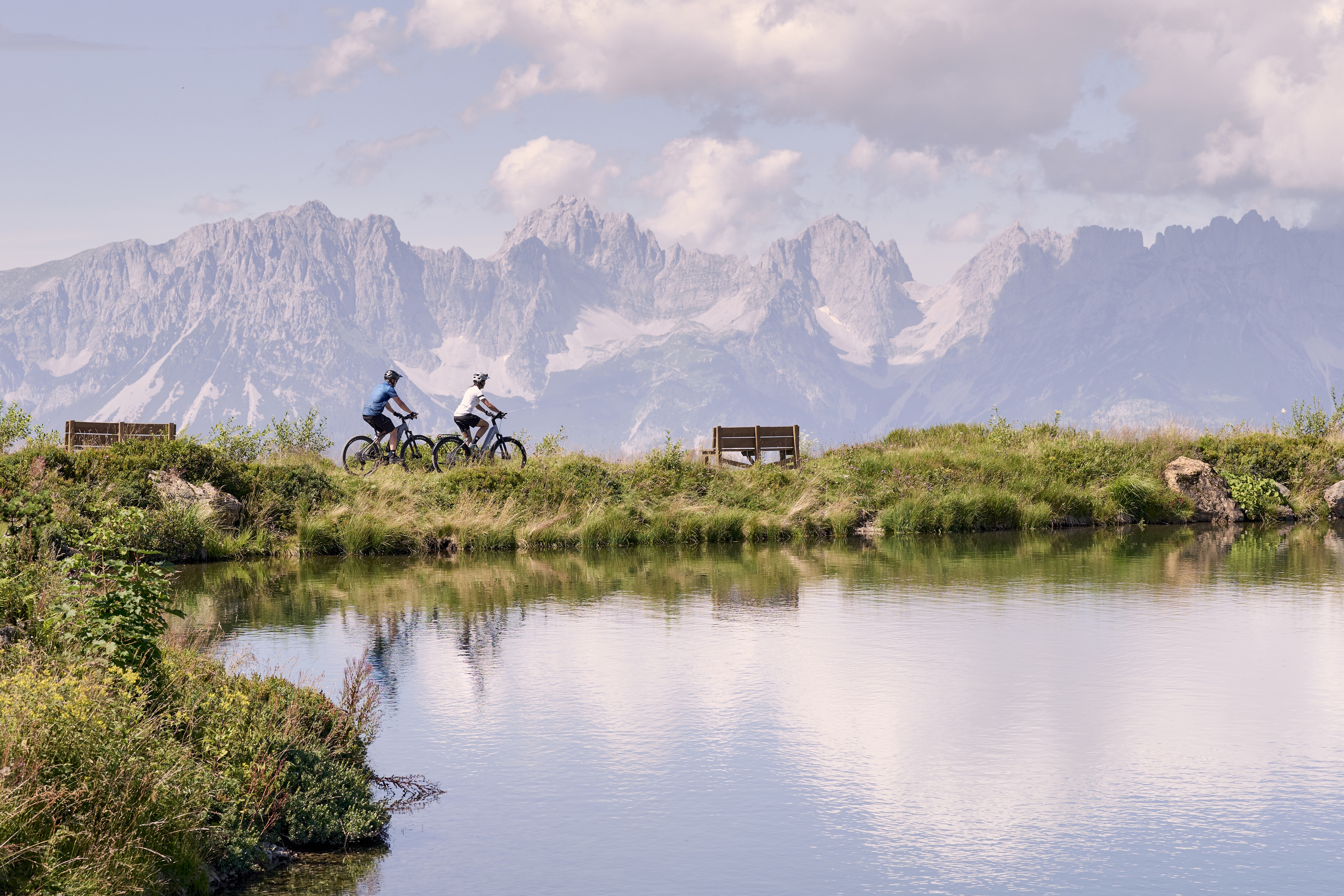 Zwei Radfahrer fahren auf einem grasbewachsenen Weg neben einem ruhigen See entlang. In der Nähe gibt es Bänke und im Hintergrund majestätische, zerklüftete Berge unter einem teilweise bewölkten Himmel.