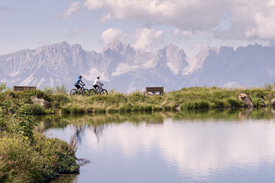 Zwei Radfahrer fahren auf einem grasbewachsenen Weg neben einem ruhigen See entlang. In der Nähe gibt es Bänke und im Hintergrund majestätische, zerklüftete Berge unter einem teilweise bewölkten Himmel.