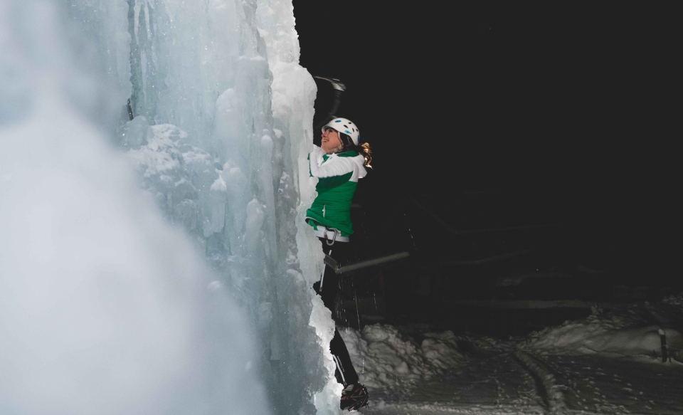 Eine Frau klettert mit Helm, Steigeisen und Eisgerät nachts an einer vereisten Wand empor.