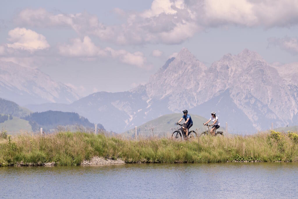 Zwei Radfahrer fahren am Seeufer entlang, mit Blick auf grüne Hügel und felsige Berggipfel im Hintergrund.