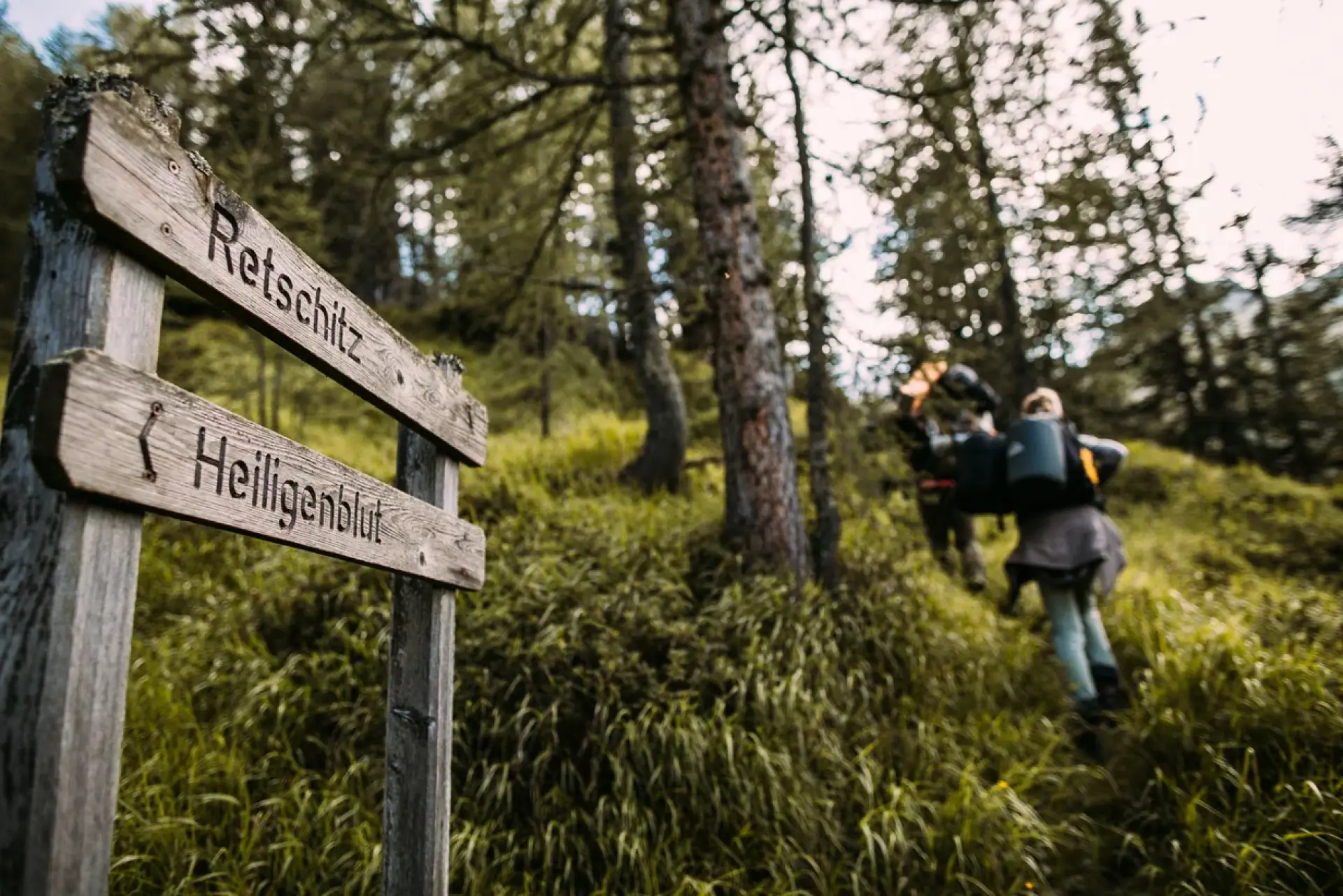 Auf einem hölzernen Wegweiser in einem Waldstück stehen die Worte „Retschitz“ und „Heiligenblut“. Im Hintergrund laufen zwei Wanderer mit Rucksäcken einen grasbewachsenen Hügel hinauf, umgeben von hohen Bäumen.