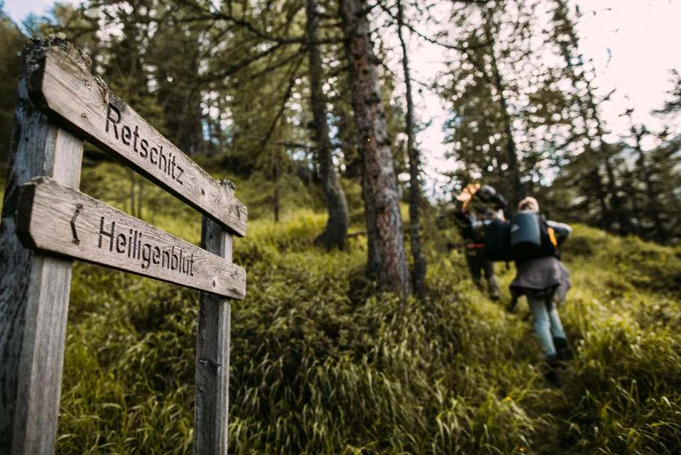 A wooden signpost in a wooded area bears the words "Retschitz" and "Heiligenblut." In the background, two hikers with backpacks are walking up a grassy hill surrounded by tall trees.