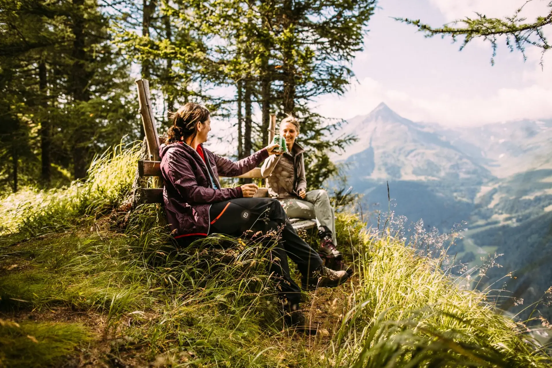 Zwei Menschen sitzen auf einem grasbewachsenen Hügel, umgeben von Bäumen, und genießen ein Getränk mit malerischer Aussicht auf die Berge im Hintergrund.