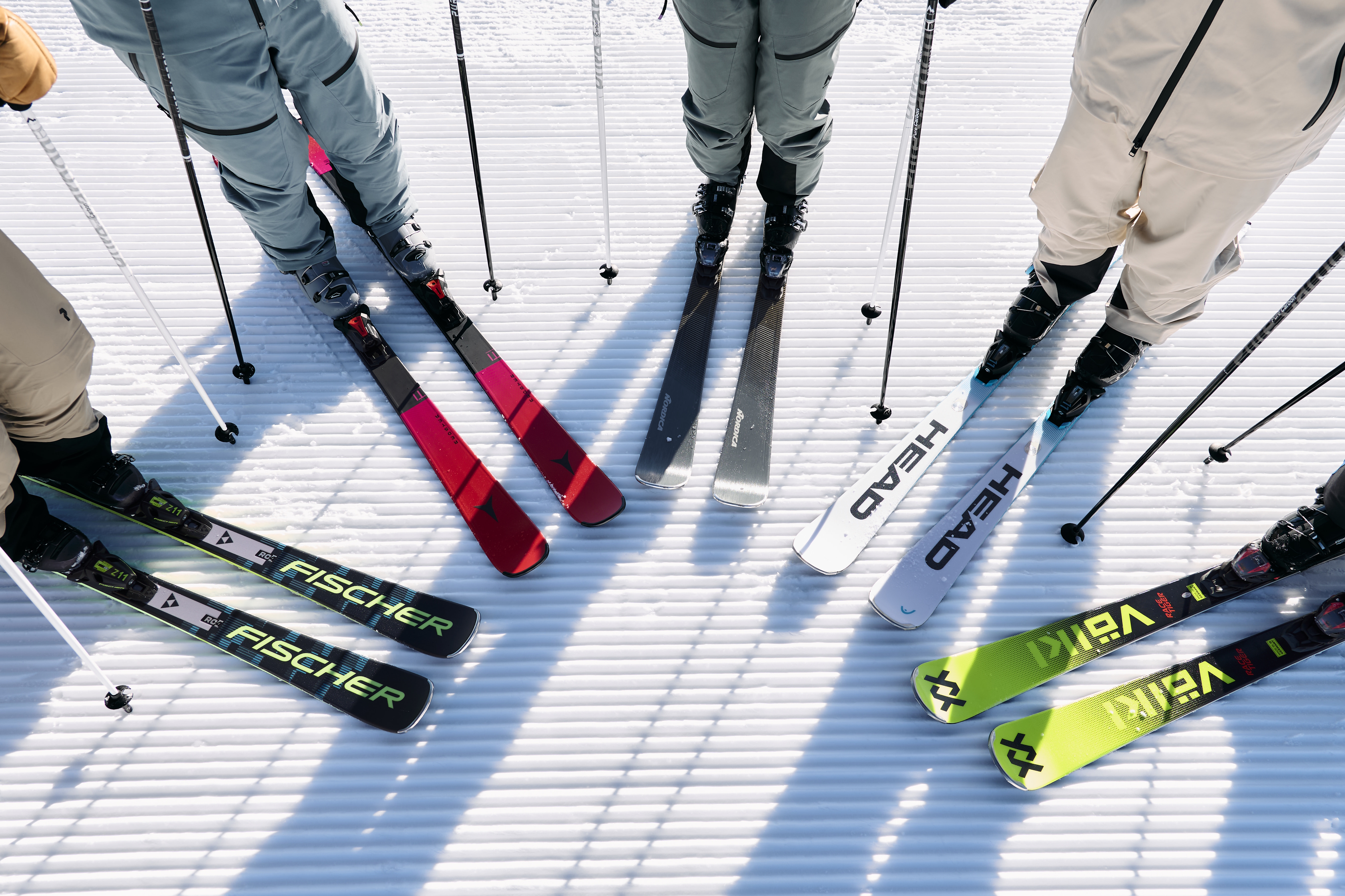 Eine Gruppe von Skifahrern steht auf einer präparierten Schneefläche. Ihre Skier und Stöcke sind im Kreis angeordnet. Die Skier sind von verschiedenen Marken und in verschiedenen Farben, darunter Rot, Schwarz, Weiß und Grün.