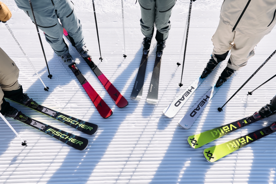 Eine Gruppe von Skifahrern steht auf einer präparierten Schneefläche. Ihre Skier und Stöcke sind im Kreis angeordnet. Die Skier sind von verschiedenen Marken und in verschiedenen Farben, darunter Rot, Schwarz, Weiß und Grün.