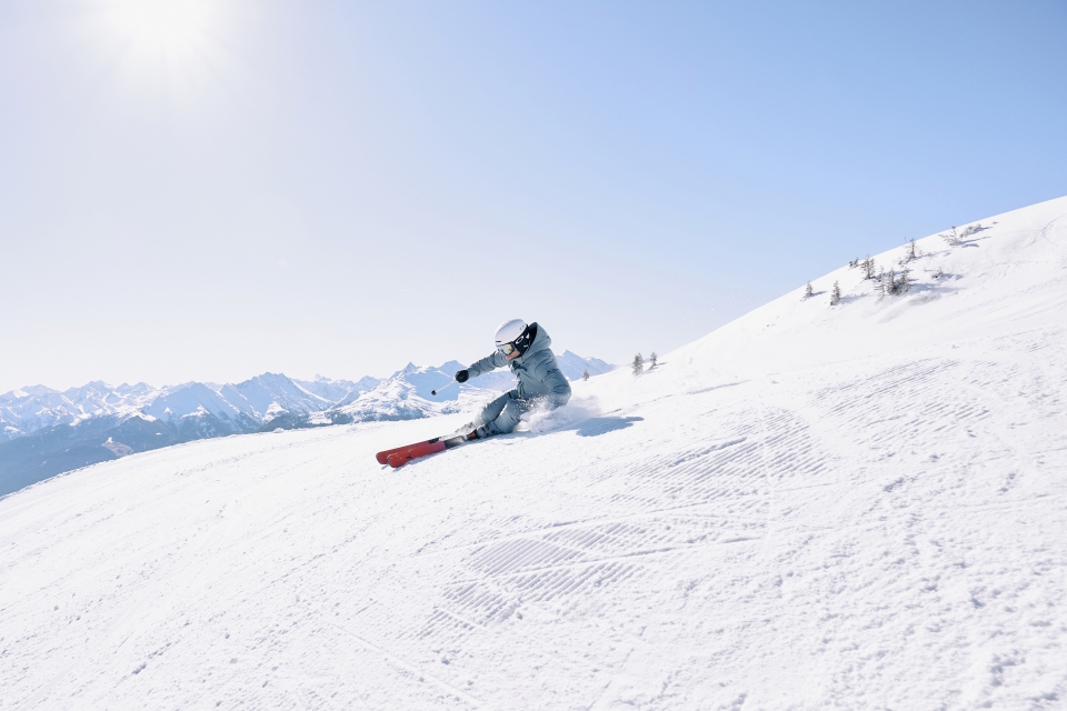 Ein Snowboarder in blauem Outfit und Helm fährt unter einem klaren blauen Himmel einen verschneiten Hang hinunter, im Hintergrund sind in der Ferne Berge zu sehen.
