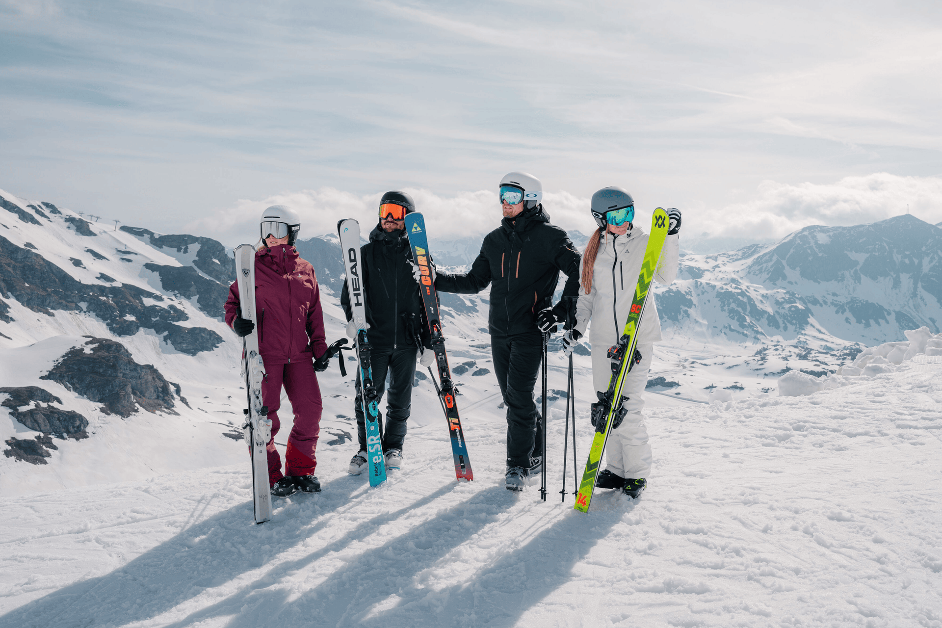 Vier Personen in Skiausrüstung stehen auf einem schneebedeckten Berghang und halten jeweils Skier in der Hand. Im Hintergrund sind schneebedeckte Gipfel unter einem teilweise bewölkten Himmel zu sehen.