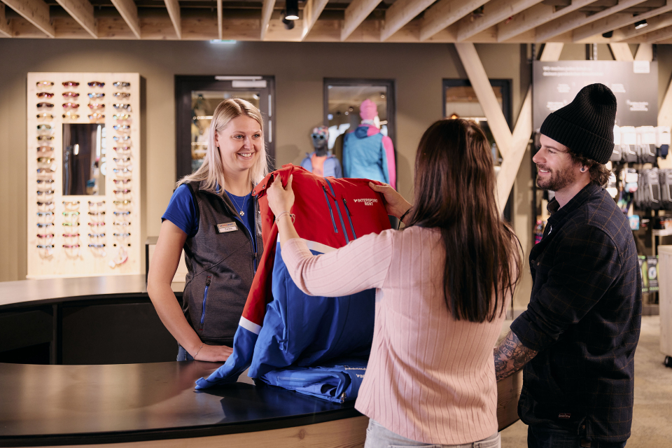 Ein Ladeninneres mit Regalen voller bunter Outdoor-Kleidung und Jacken auf beiden Seiten. In der Mitte befindet sich ein Ausstellungstisch mit gefalteter Kleidung und Schuhen. Das Geschäft ist modern gestaltet, mit Teppichboden und einer Theke im Hintergrund.