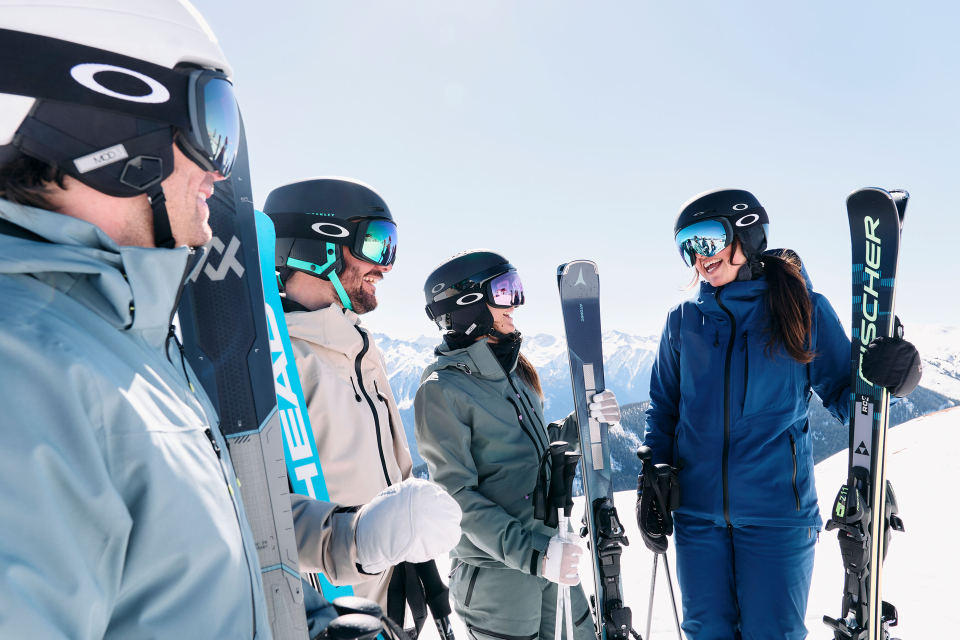 Eine Gruppe von vier Personen in Skiausrüstung, einschließlich Helmen und Schutzbrillen, steht mit Skiern in der Hand an einem schneebedeckten Berghang. Sie lächeln und unterhalten sich, im Hintergrund ein strahlend blauer Himmel und in der Ferne schneebedeckte Berge.