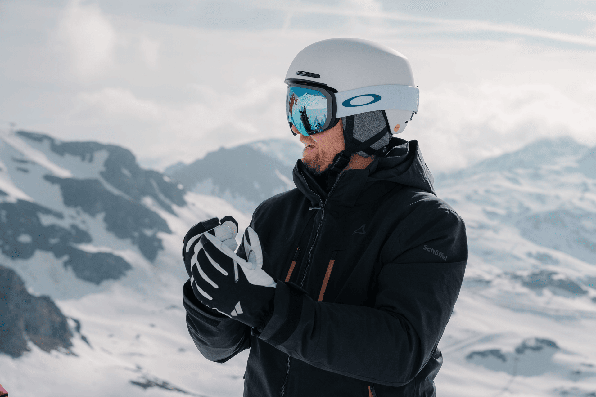 Eine Person mit weißem Skihelm und reflektierender Schutzbrille steht in einer verschneiten Berglandschaft. Sie trägt eine schwarze Jacke und Handschuhe. Im Hintergrund sind schneebedeckte Gipfel unter einem teilweise bewölkten Himmel zu sehen.