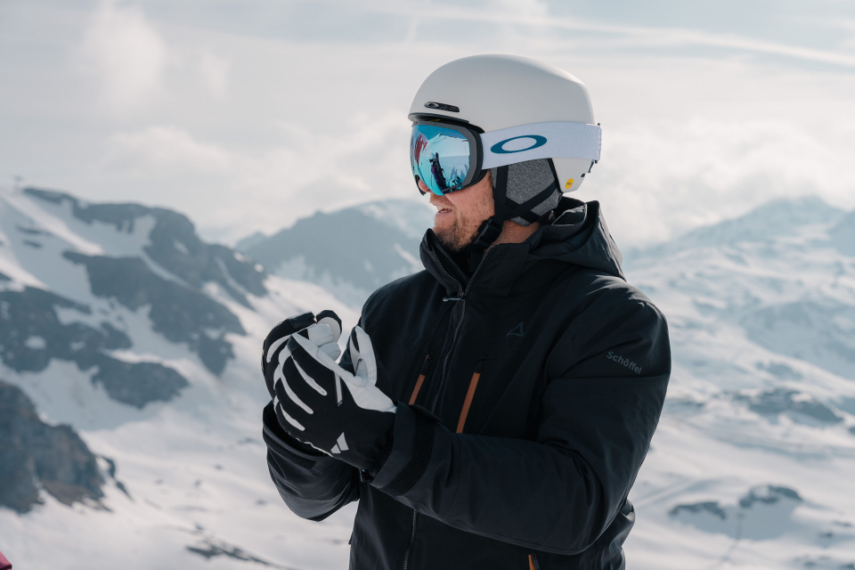 Eine Person mit weißem Skihelm und reflektierender Schutzbrille steht in einer verschneiten Berglandschaft. Sie trägt eine schwarze Jacke und Handschuhe. Im Hintergrund sind schneebedeckte Gipfel unter einem teilweise bewölkten Himmel zu sehen.