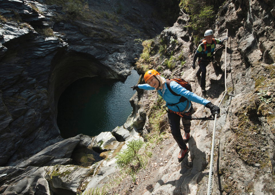 Two people wearing helmets and climbing gear navigate a steep, rocky path beside a deep, circular rock formation with water at the bottom. The person in front is smiling and gesturing outward, while the person behind follows closely.